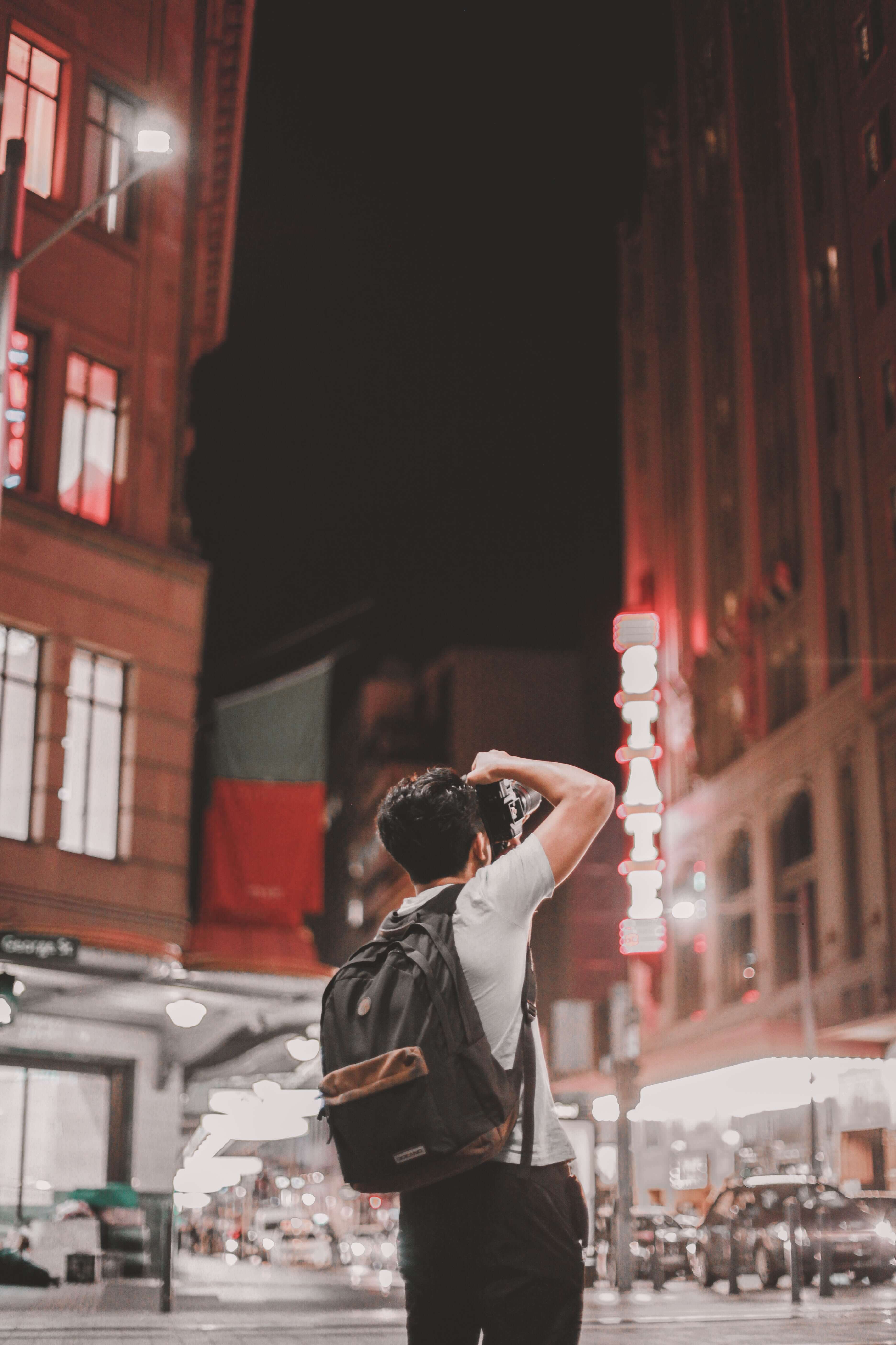 Man in white top carrying backpack holding black DSLR camera outdoors